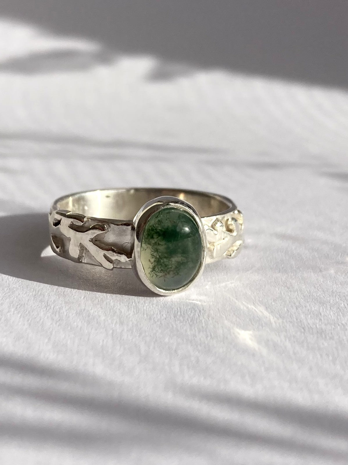 A sterling silver ring with a textured, organic-shaped band and a central oval cabochon moss agate green stone, resting on a white textured surface with soft shadows.