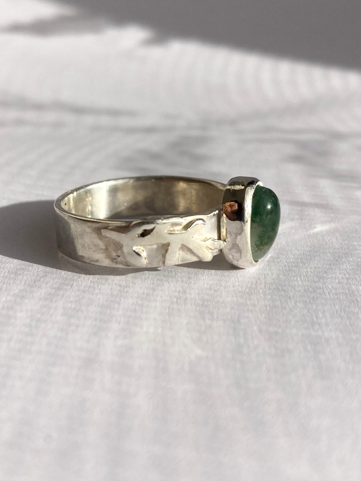 A silver ring with leaf detail band and a bezel-set oval green moss agate stone, resting on a white textured fabric surface.