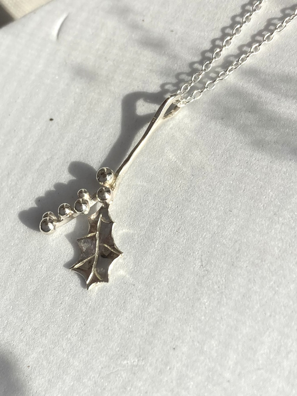 A sterling silver holly leaf pendant necklace on a delicate link chain, displayed on a textured white surface with soft leaf shadows.