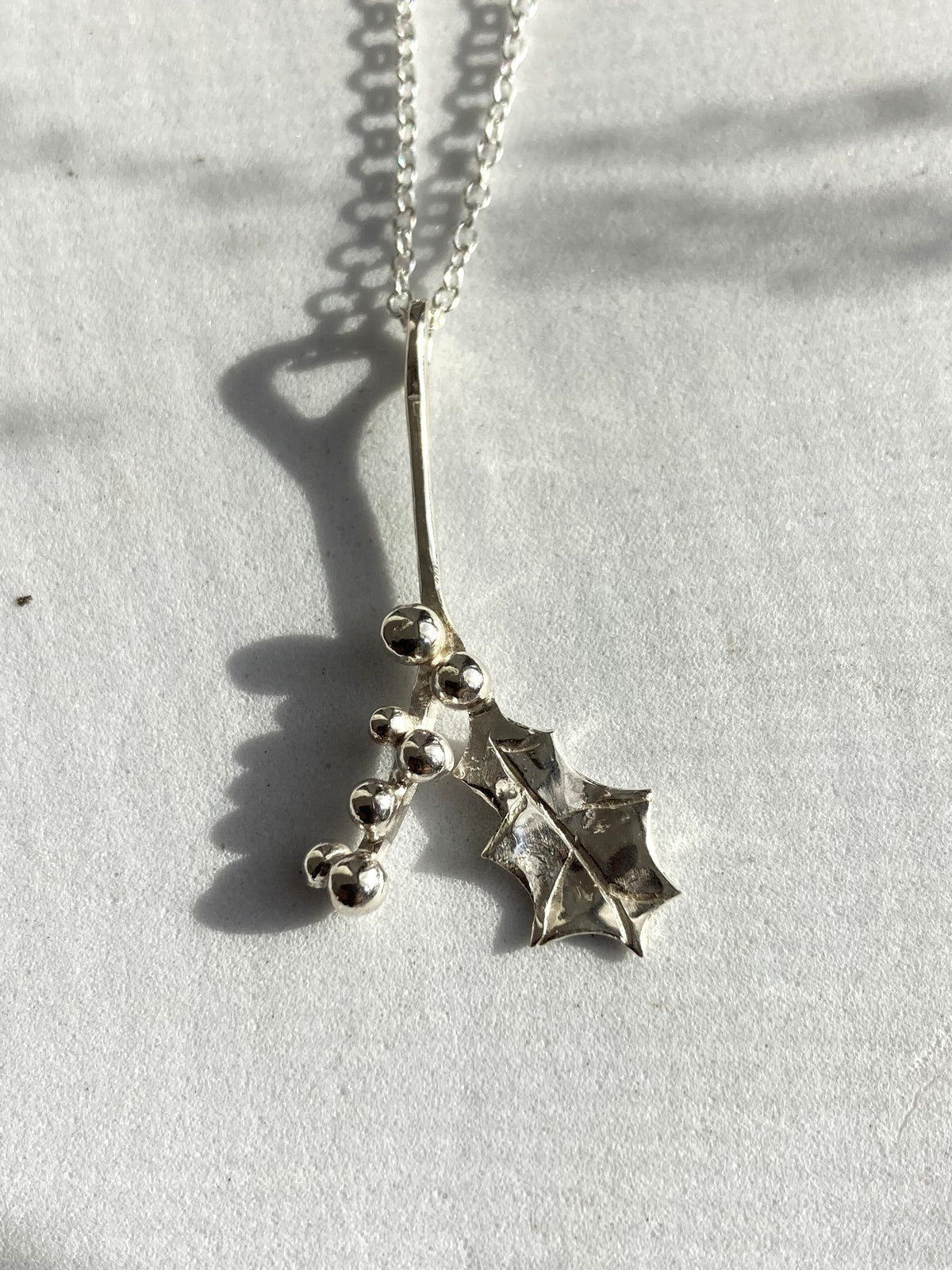 A silver holly and berry pendant necklace on a delicate silver chain, photographed on a textured white surface with strong natural shadows.