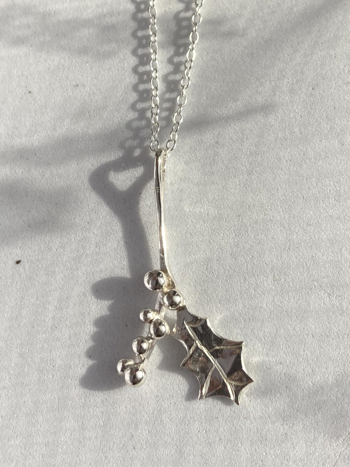 A sterling silver pendant necklace featuring a holly leaf and small silver berries, displayed on a textured light grey surface with natural shadows.