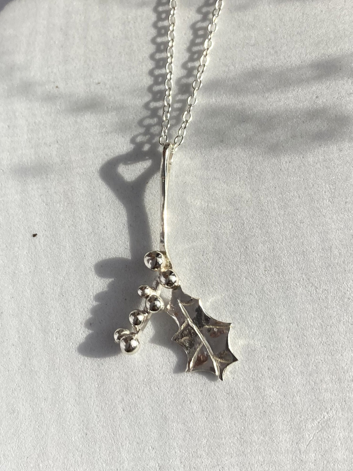 A sterling silver necklace with a holly leaf and berry pendant, photographed on a light textured surface with strong natural shadows.
