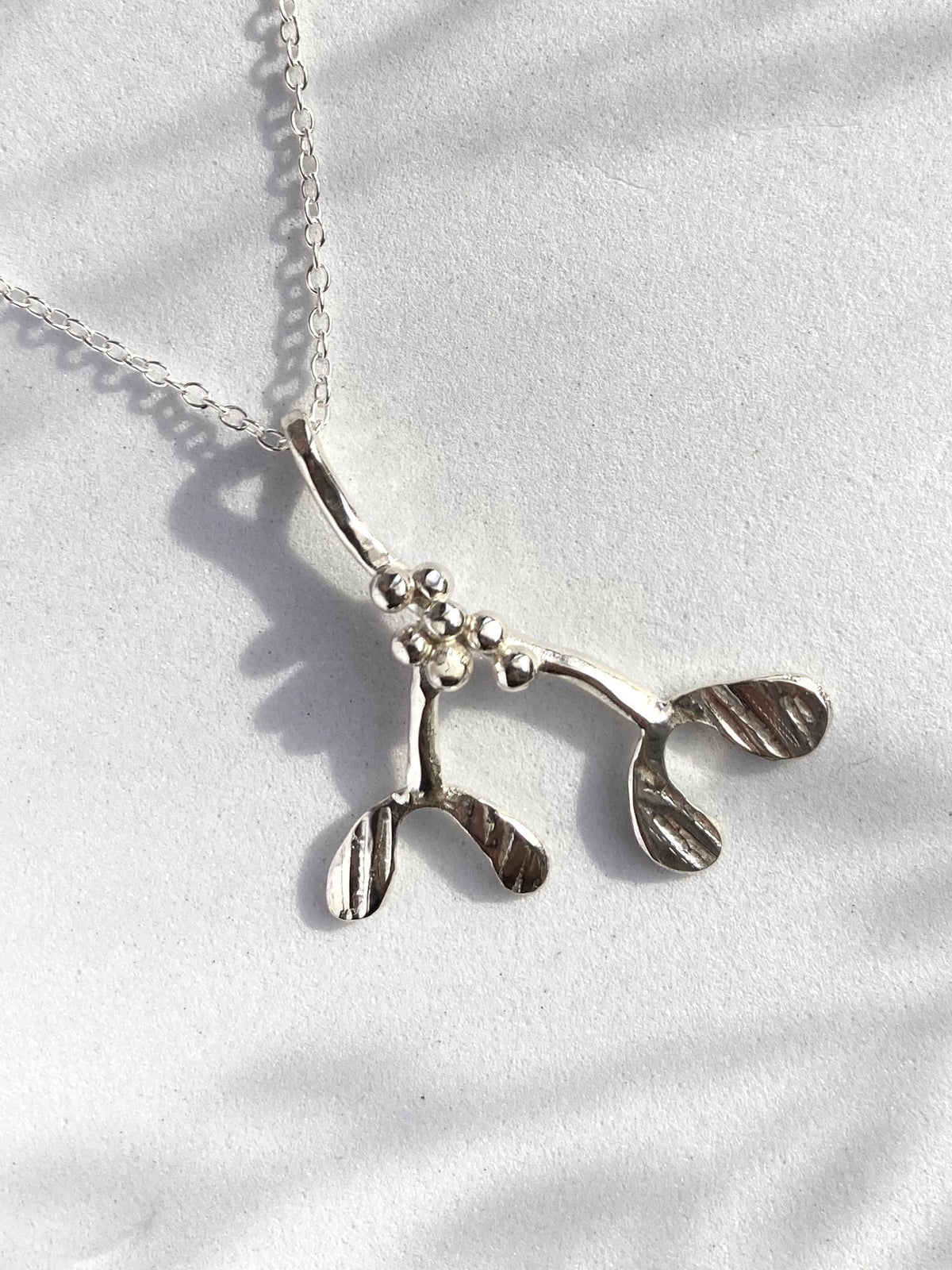 A sterling silver mistletoe pendant necklace on a delicate link chain, displayed on a textured white fabric background with soft shadows.