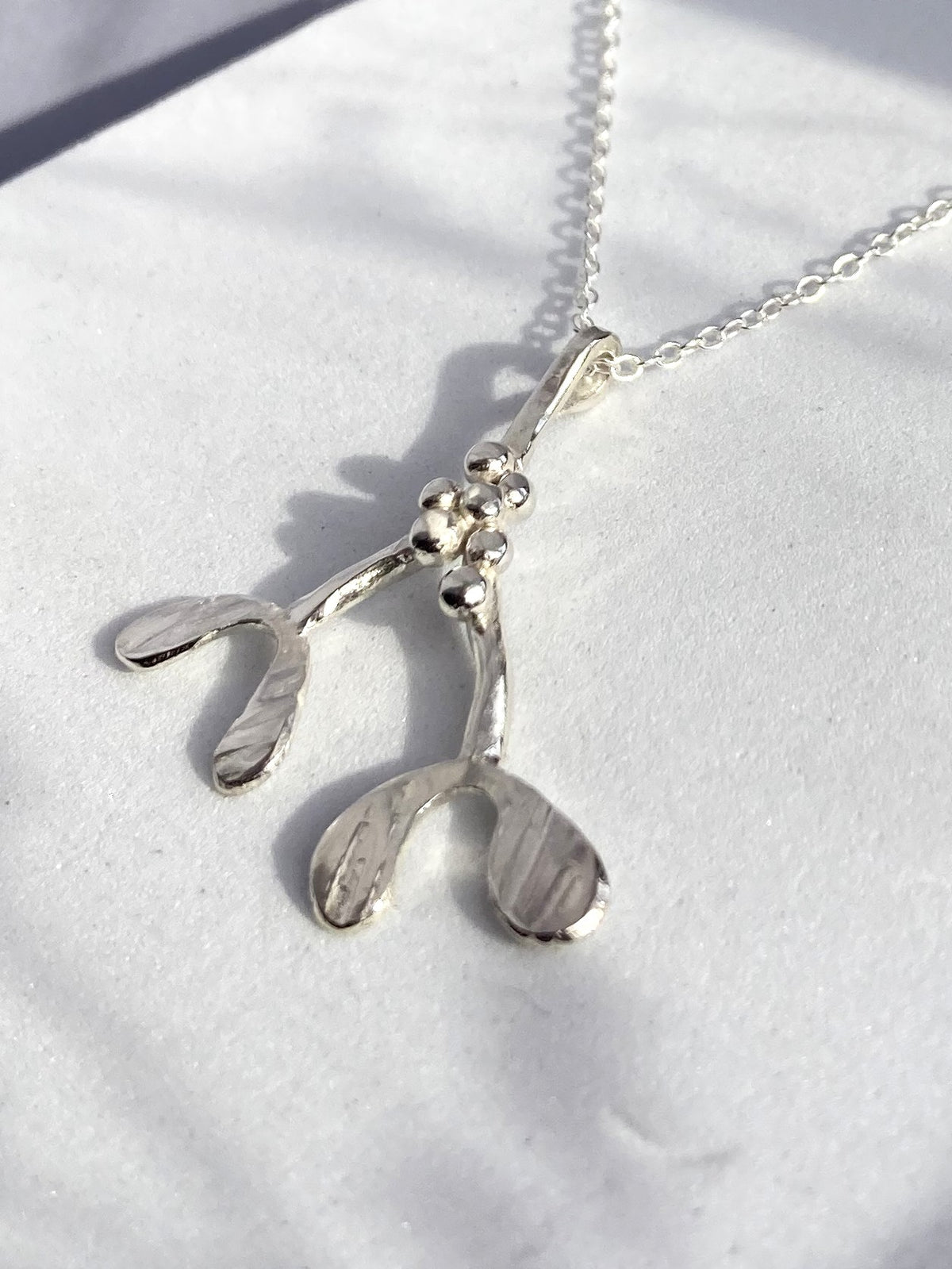 A sterling silver mistletoe pendant necklace on a delicate chain, displayed on a textured white surface with soft shadows.