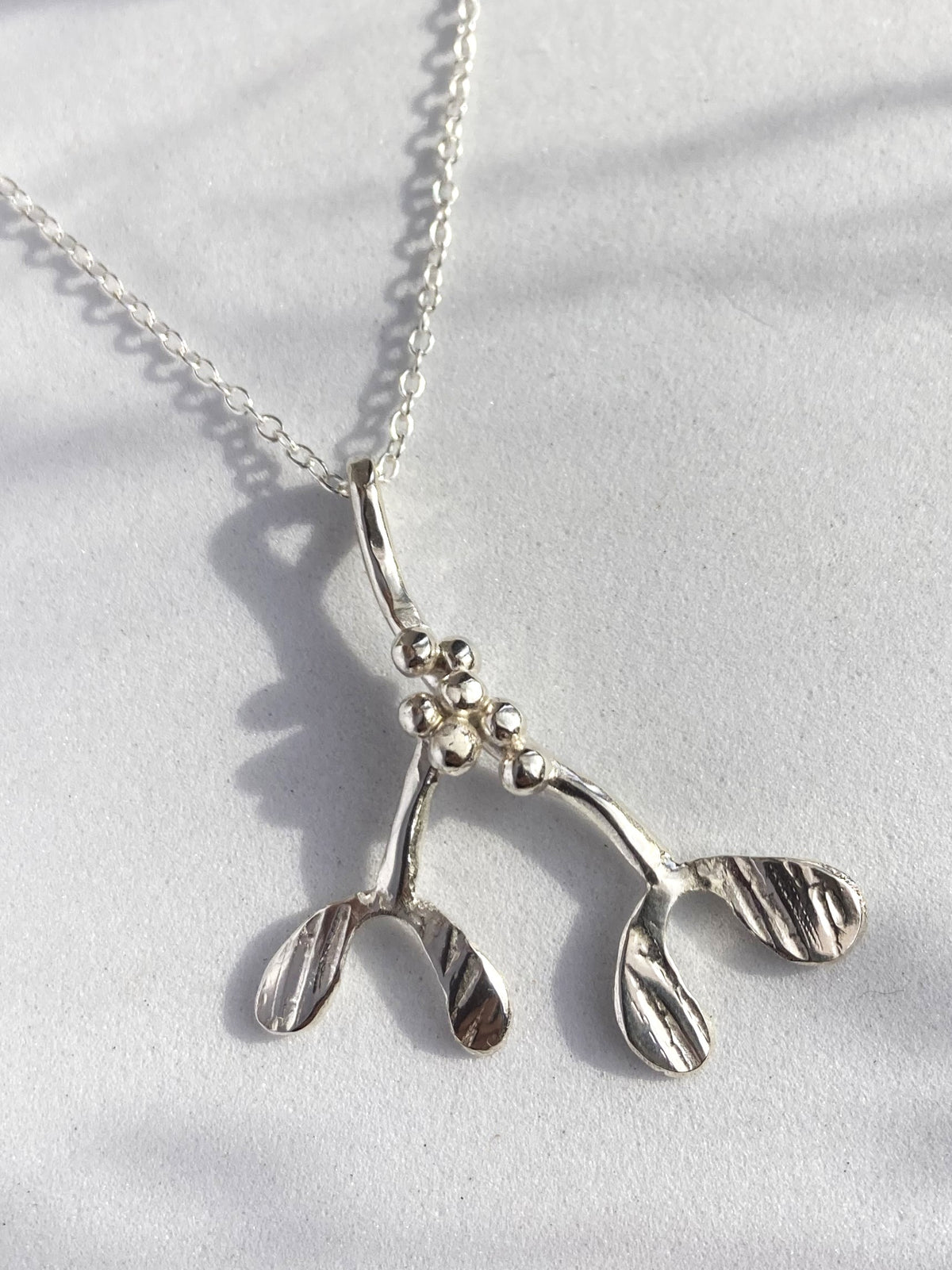 A textured sterling silver mistletoe pendant necklace on a delicate link chain, displayed on a textured white surface with soft shadows.