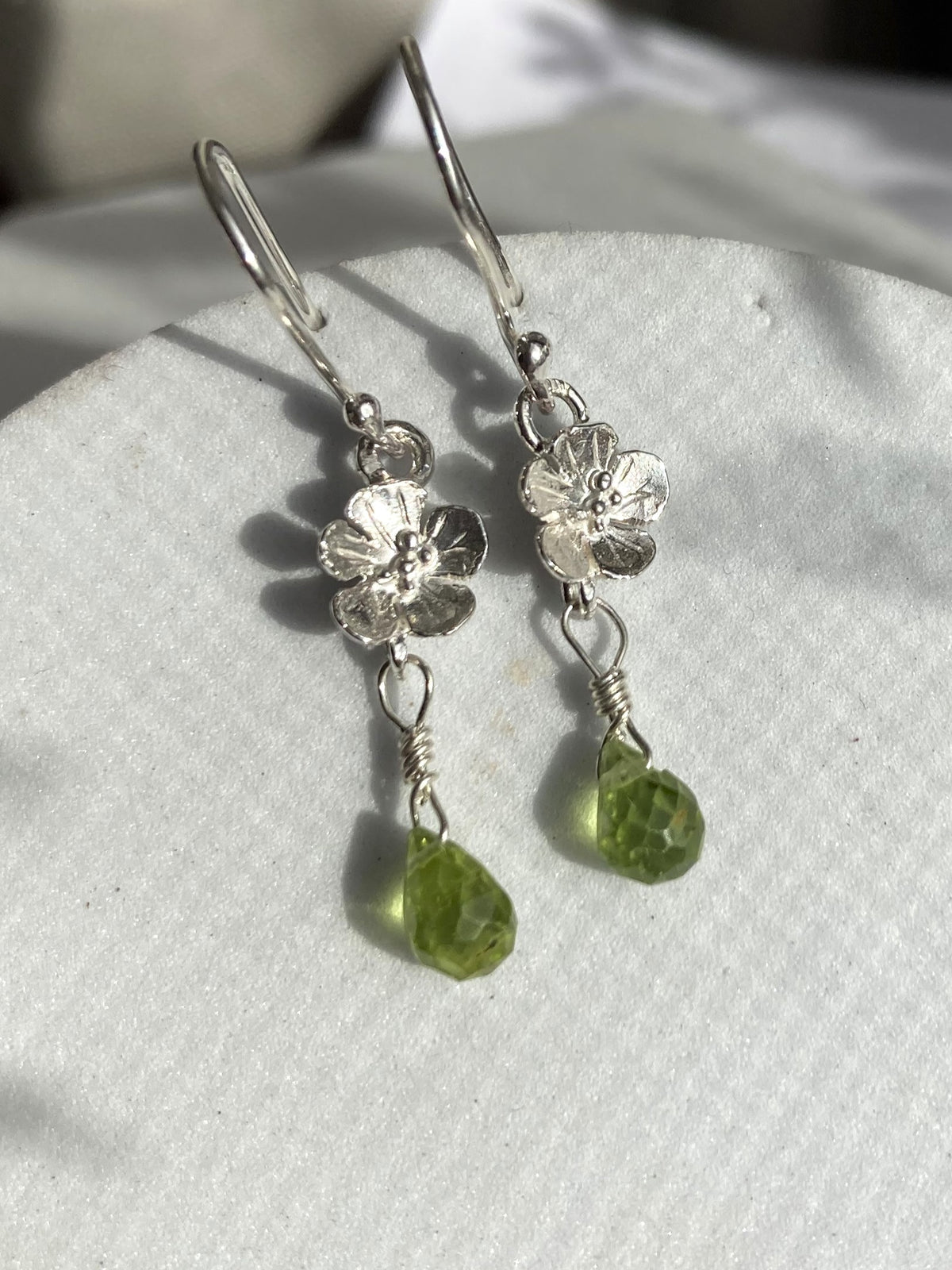 A pair of silver dangle earrings with textured flower charms and faceted green peridot gemstone drops, displayed on a textured white surface.
