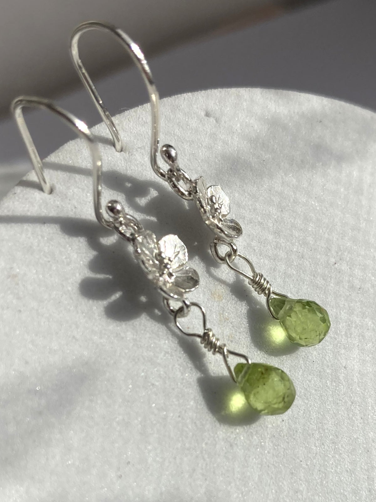 A pair of silver dangle earrings with textured silver charms and faceted light green briolette, displayed on a white textured surface.