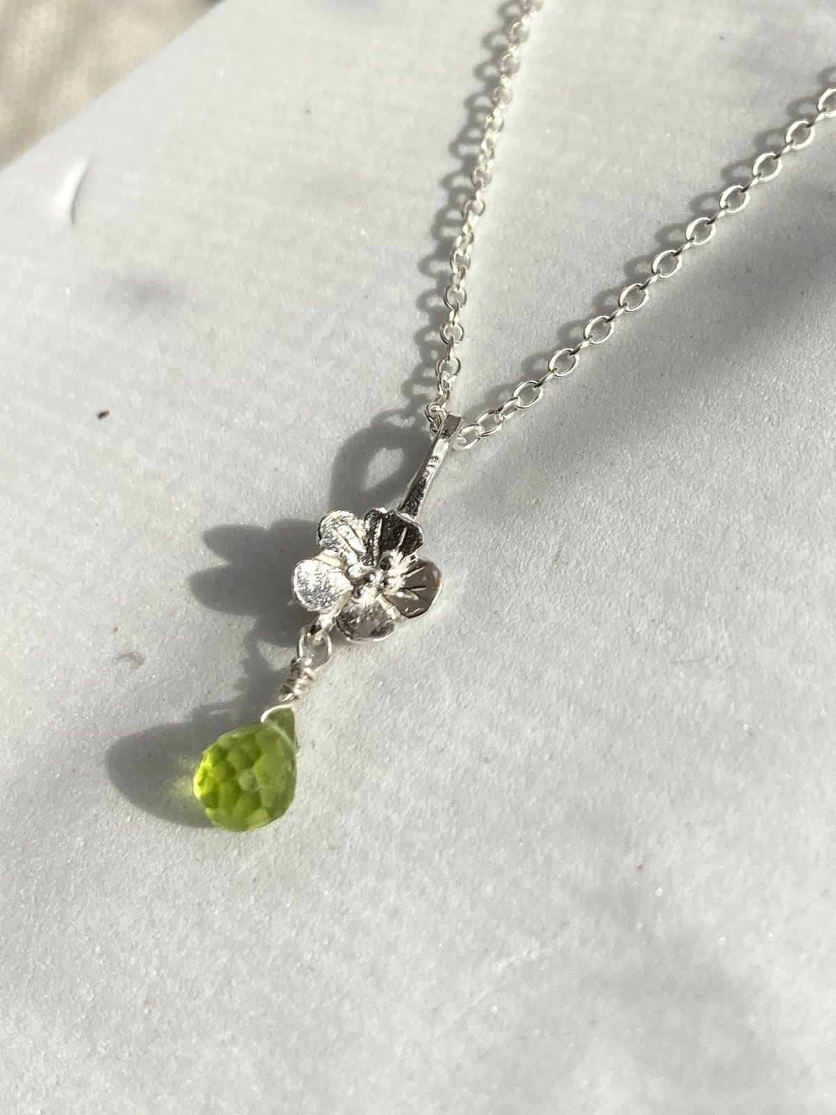 A silver flower pendant necklace with a faceted green teardrop peridot gemstone, displayed on a textured white surface.