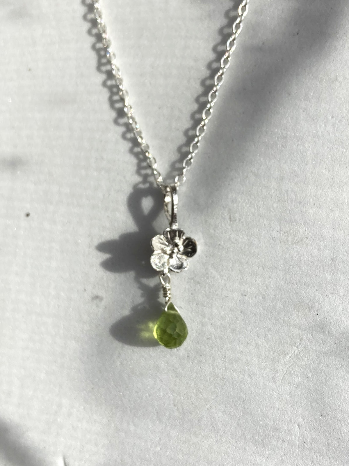 A silver chain necklace with a small flower-shaped silver pendant and a dangling green teardrop gemstone, shown on a light gray textured surface with shadows.