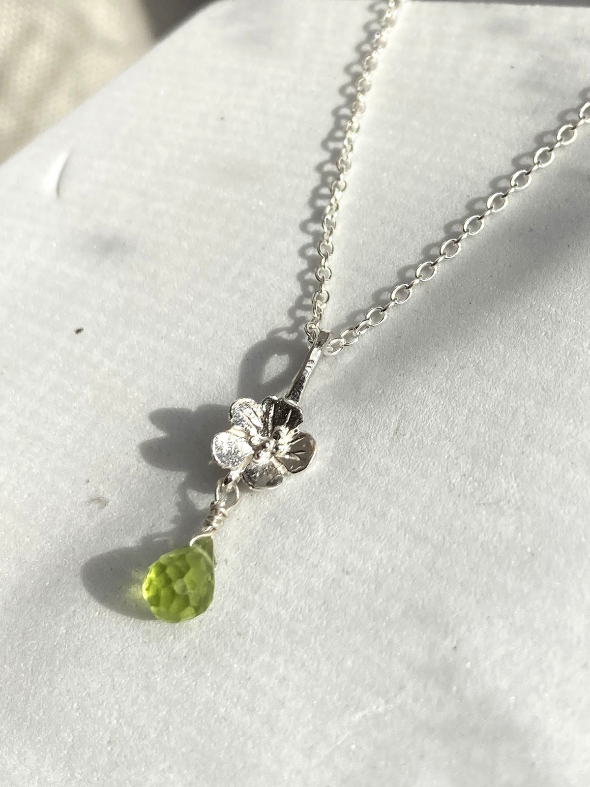 A silver necklace with a small flower-shaped pendant and a faceted green peridot briolette gemstone drop, displayed on a textured white surface.