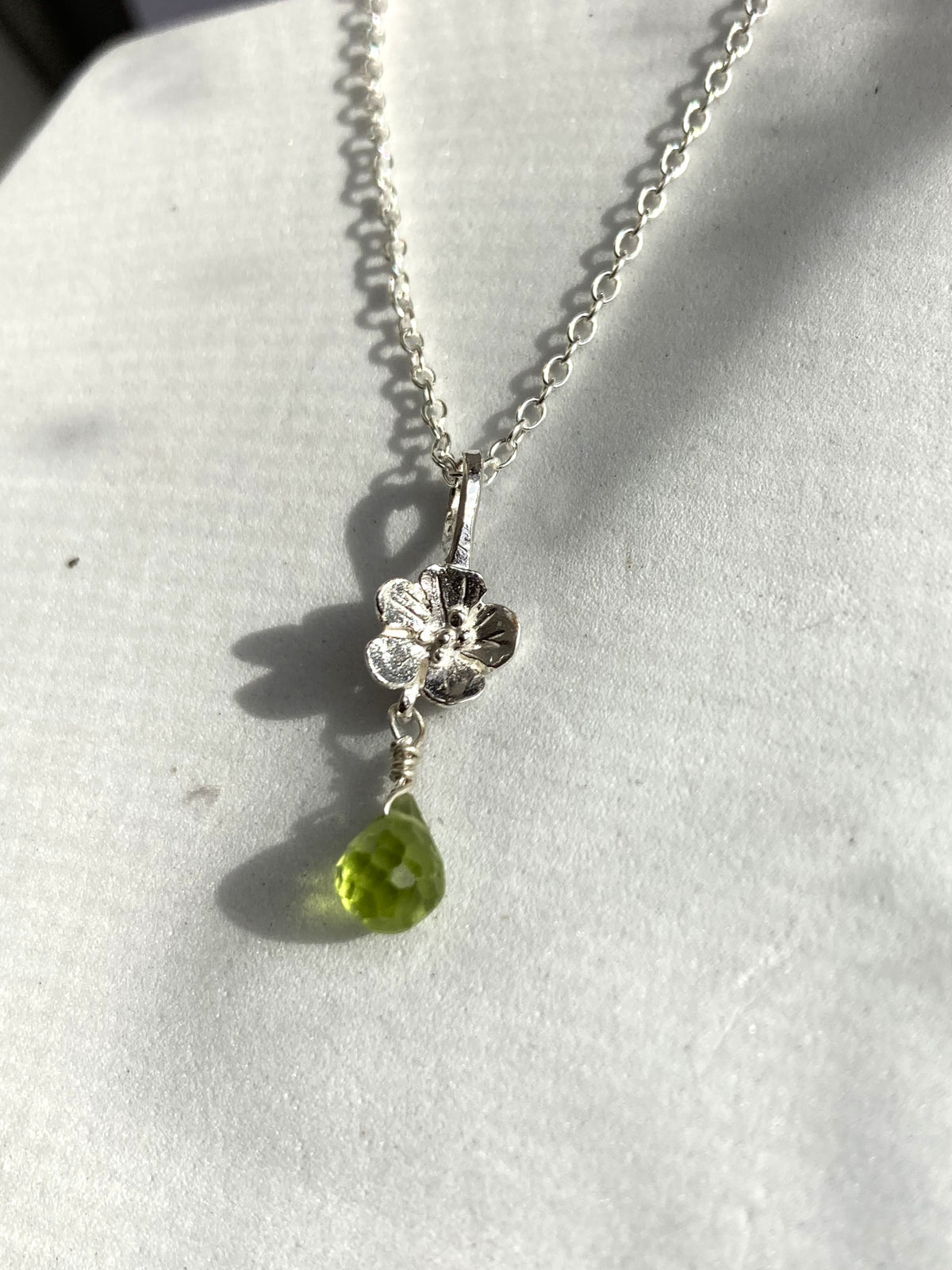 A silver chain necklace with a silver flower pendant and a dangling green teardrop gemstone, displayed on a white textured surface.