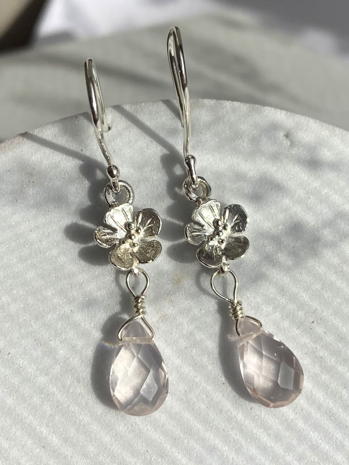 A pair of silver drop earrings by Kim Styles Jewellery, featuring textured flower charms and faceted pale pink teardrop gemstones, displayed on a textured white surface.