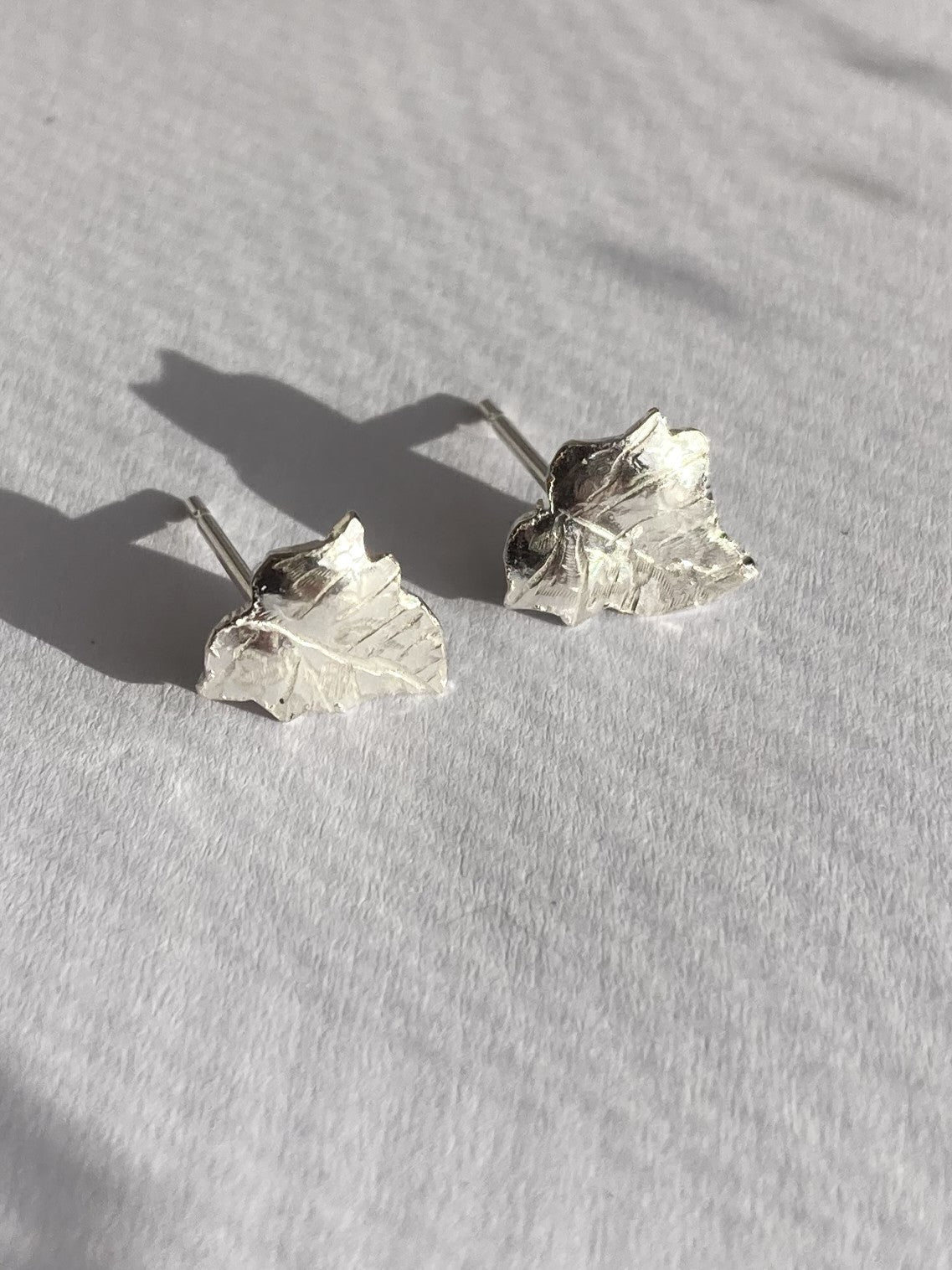 A pair of silver Ivy leaf stud earrings with textured, hammered details, resting on a white textured fabric surface with soft shadows.