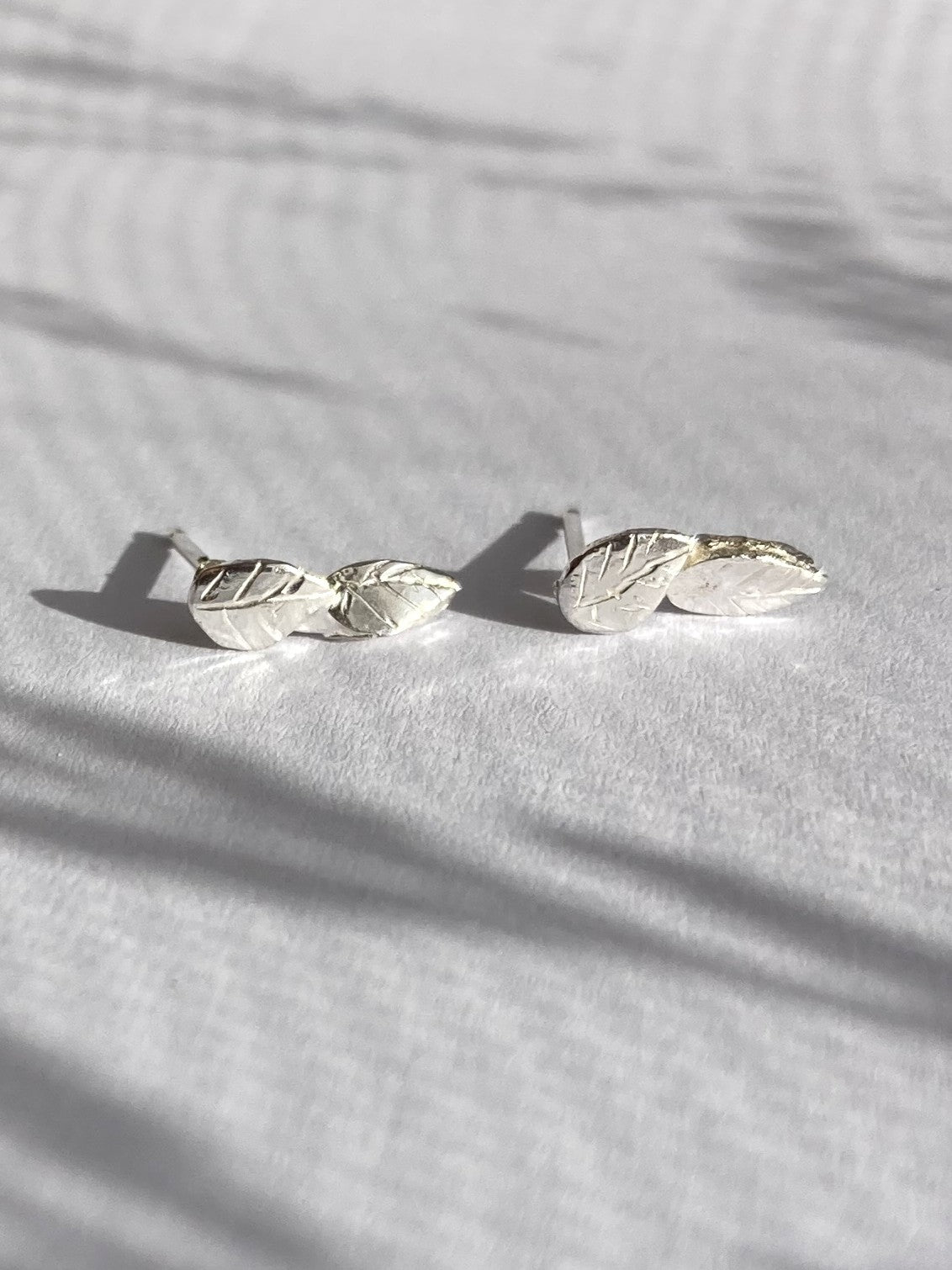 A pair of silver leaf stud earrings resting on a textured white surface with soft palm leaf shadows cast across them.