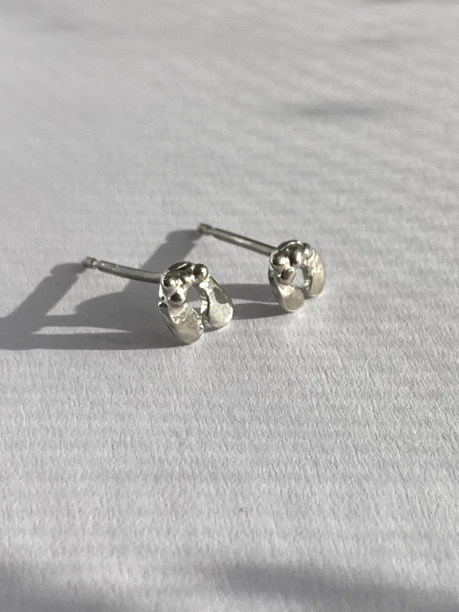 A pair of small silver stud earrings with textured, organic mistletoe leaves, resting on a white textured fabric surface with soft shadows.