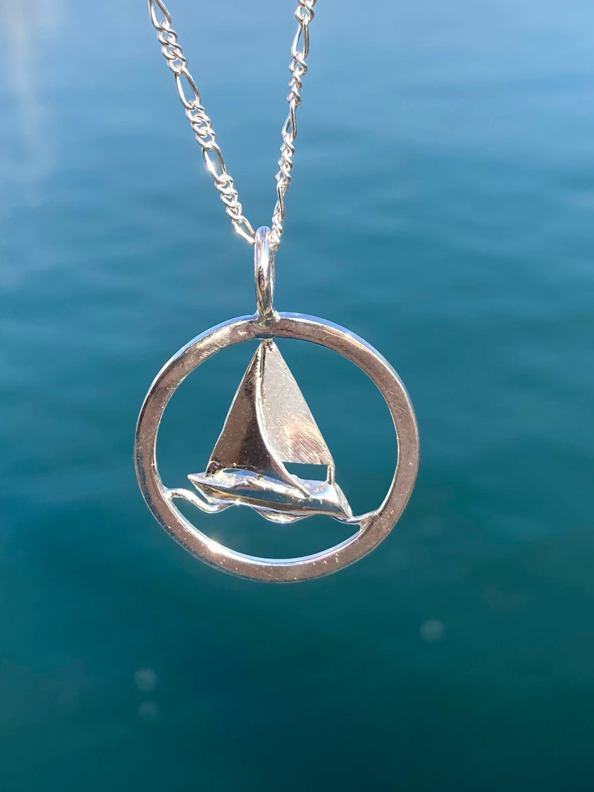 A silver pendant necklace featuring a small sailboat inside an open circular frame, hanging from a silver chain against a blurred blue water background.