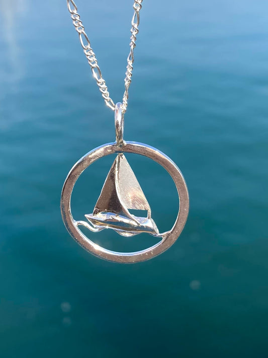 A silver pendant necklace featuring a small sailboat inside an open circular frame, hanging from a silver chain against a blurred blue water background.