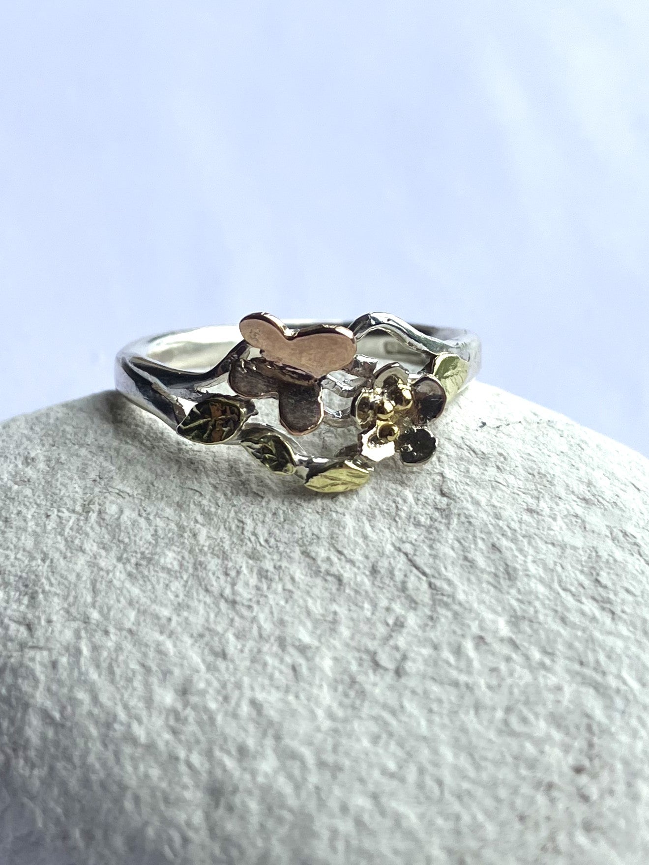 Handmade silver and gold butterfly garden ring with white gold flower, rose gold butterfly and green gold leaves, one-of-a-kind woodland inspired asymmetrical ring by Kim Styles, on white pebble