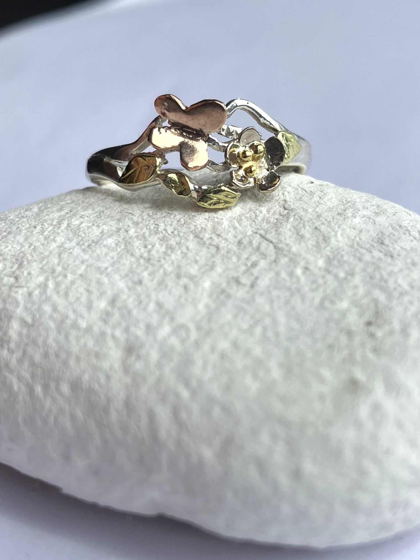Handmade silver and gold butterfly garden ring with white gold flower, rose gold butterfly and green gold leaves, one-of-a-kind woodland inspired asymmetrical ring, on white pebble
