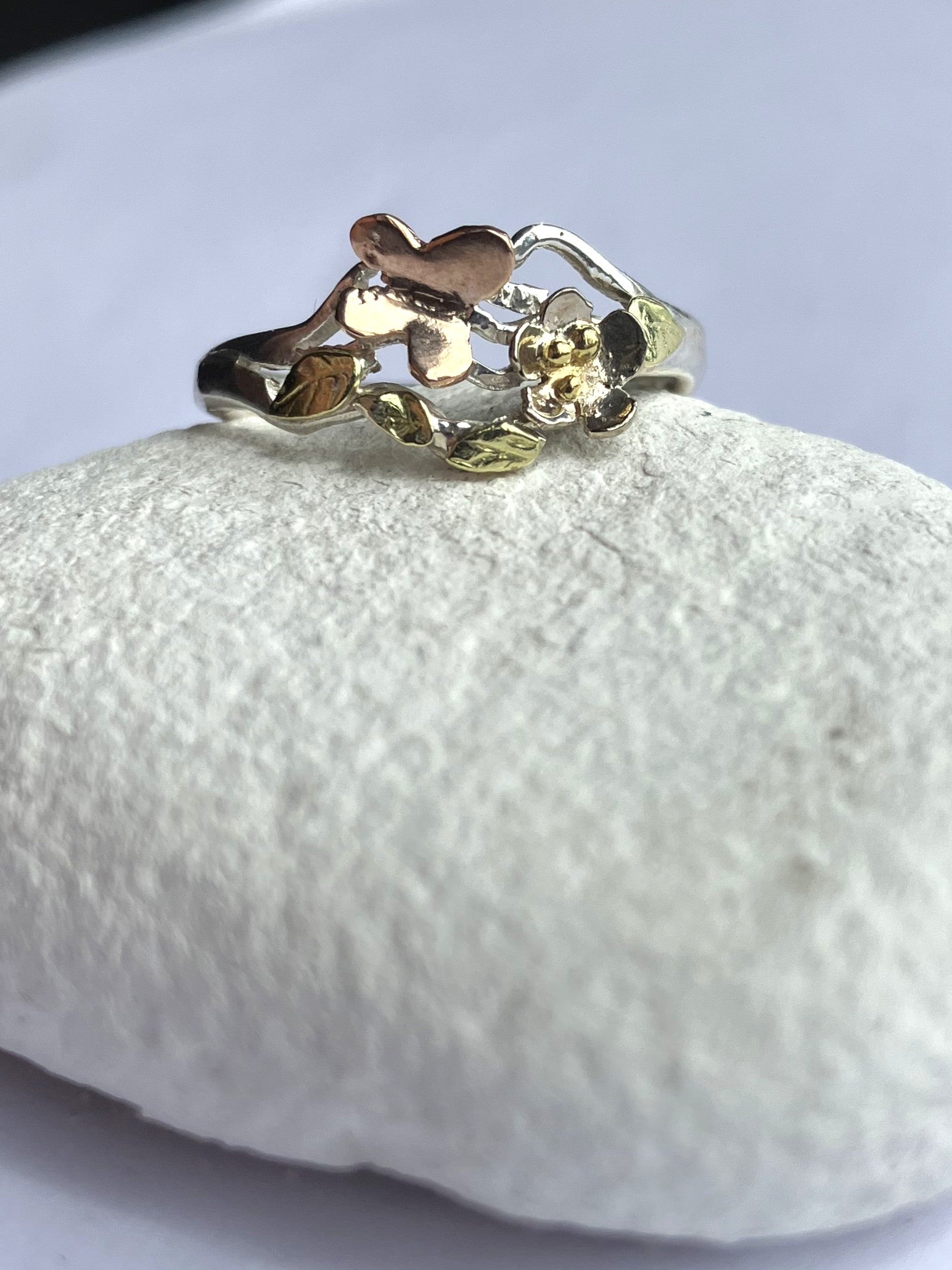 Handmade silver and gold butterfly garden ring with white gold flower, rose gold butterfly and green gold leaves, one-of-a-kind woodland inspired asymmetrical ring, on white pebble
