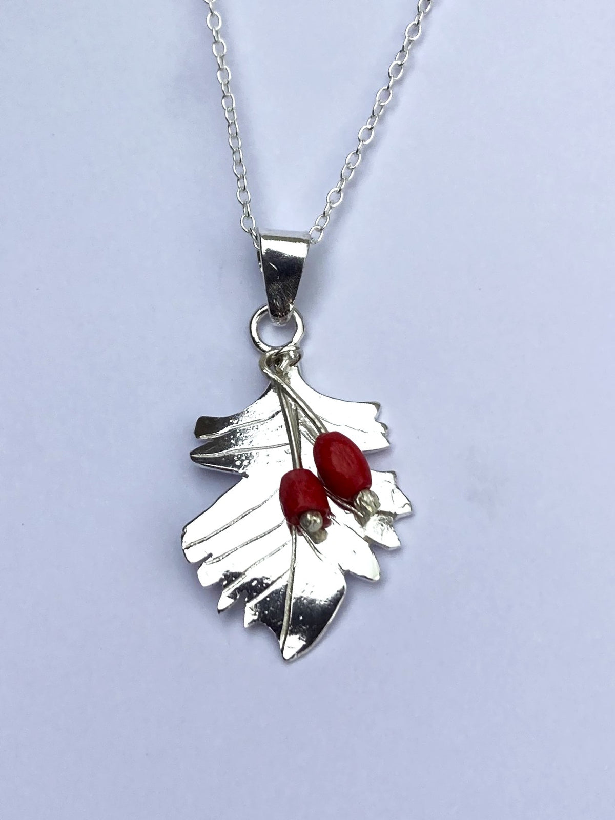 A handmade silver leaf pendant necklace featuring two small red berry accents, shown on a grey background.