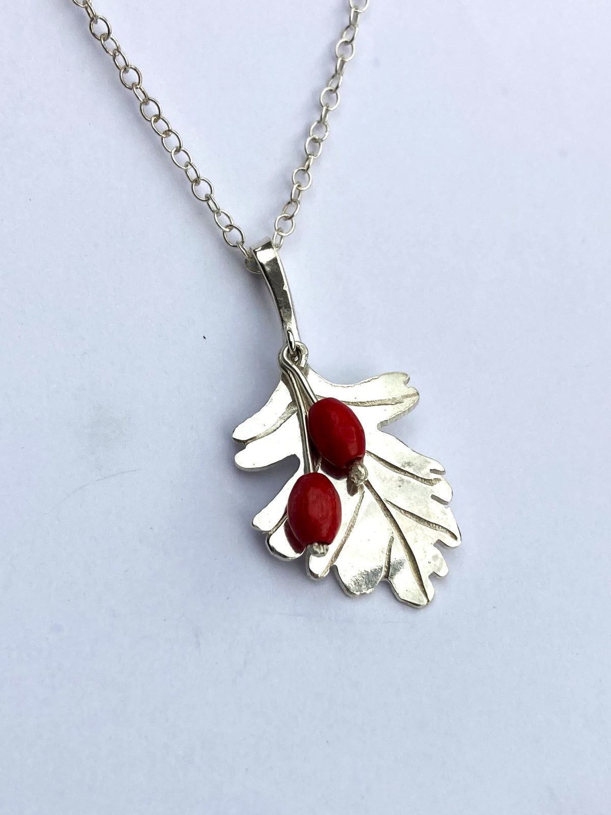 A silver pendant necklace by Kim Styles Jewellery featuring a leaf design with two small red berry accents, shown on a light grey background.