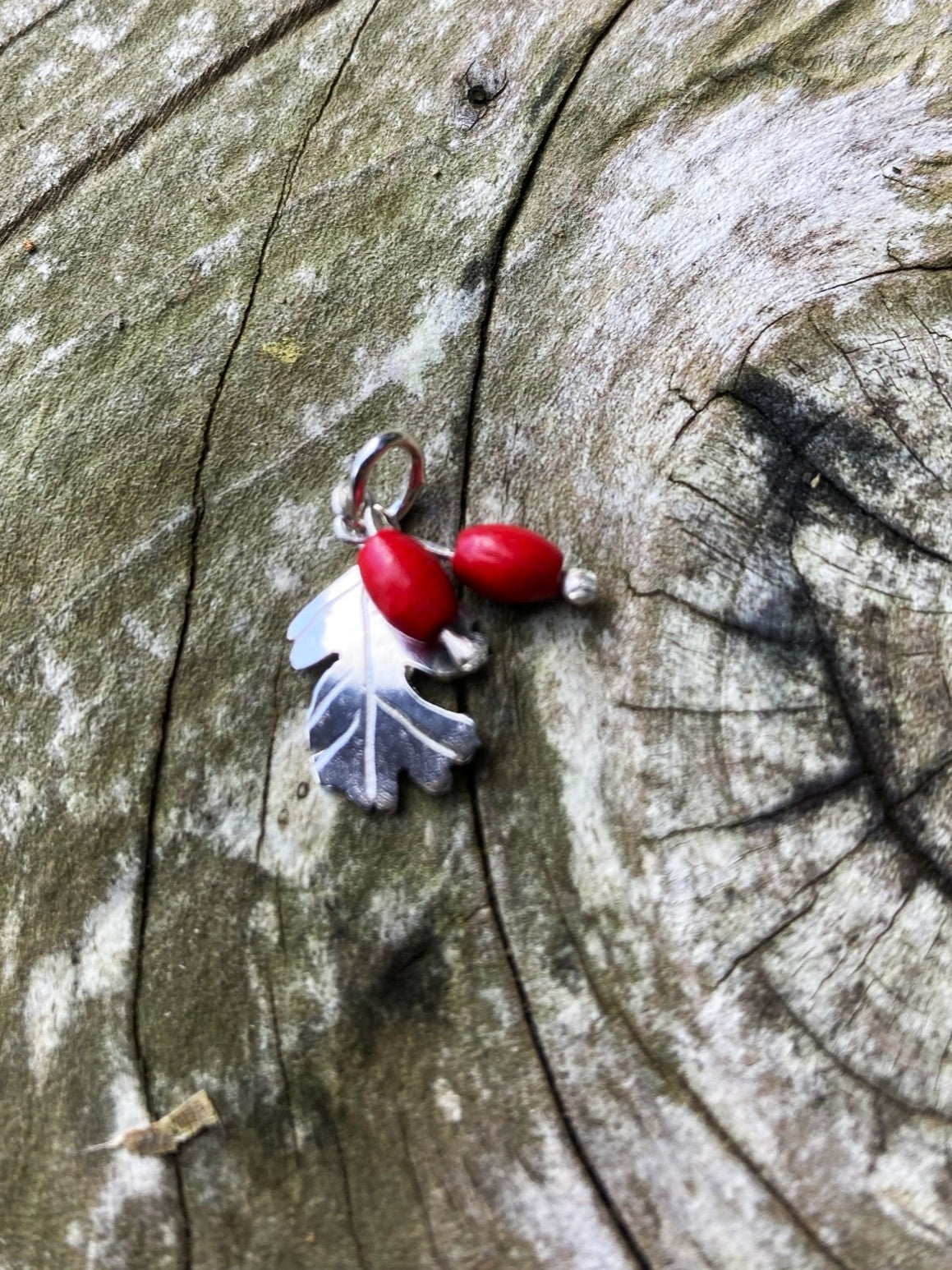 A silver Hawthorne leaf pendant charm with red beads, resting on a textured, weathered wood surface.