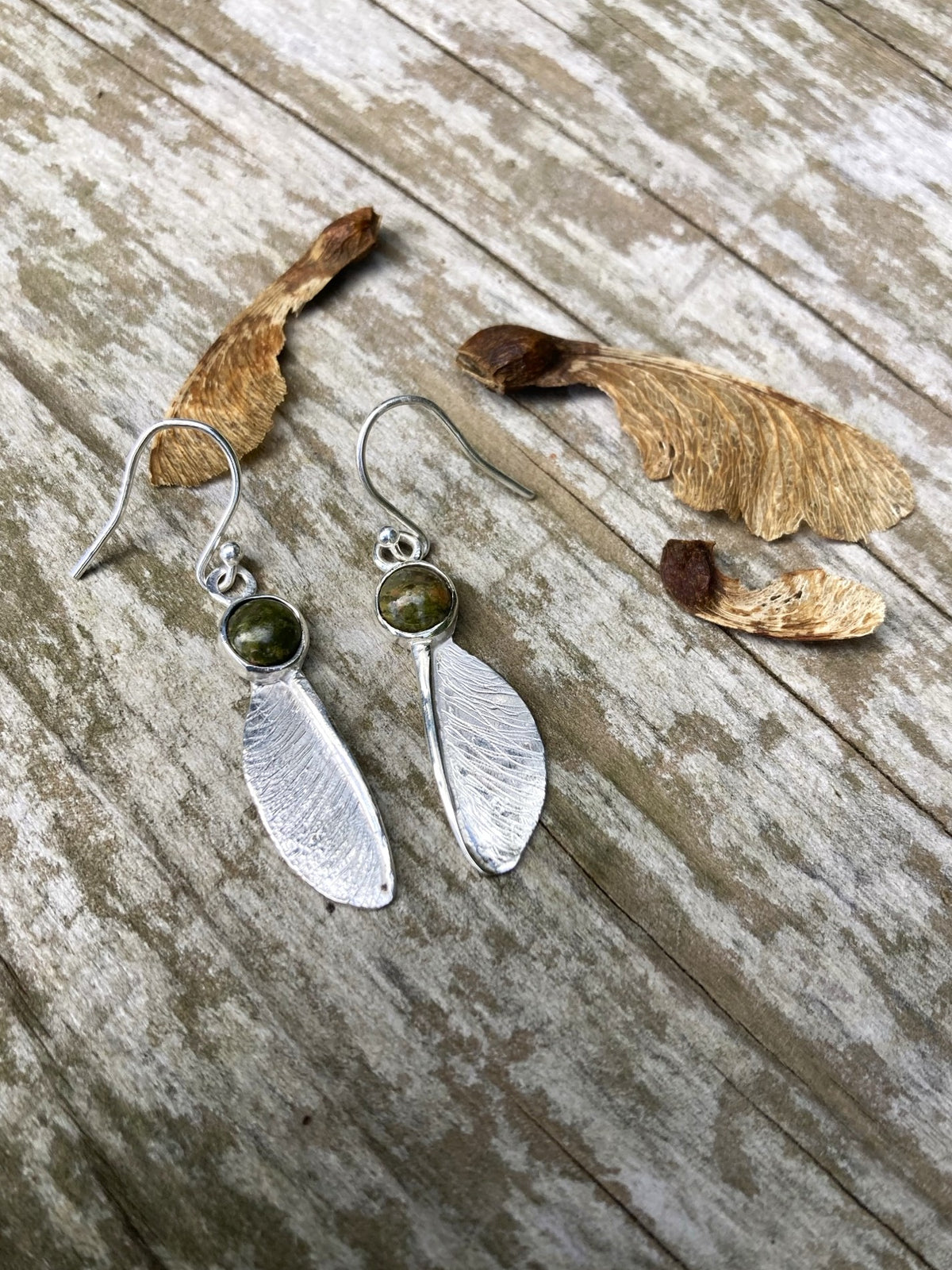 Pair of hook earrings in sterling silver  by Kim Styles Jewellery with sycamore seed design set with round cabochon green agates, on textured wood background with real sycamore seeds 