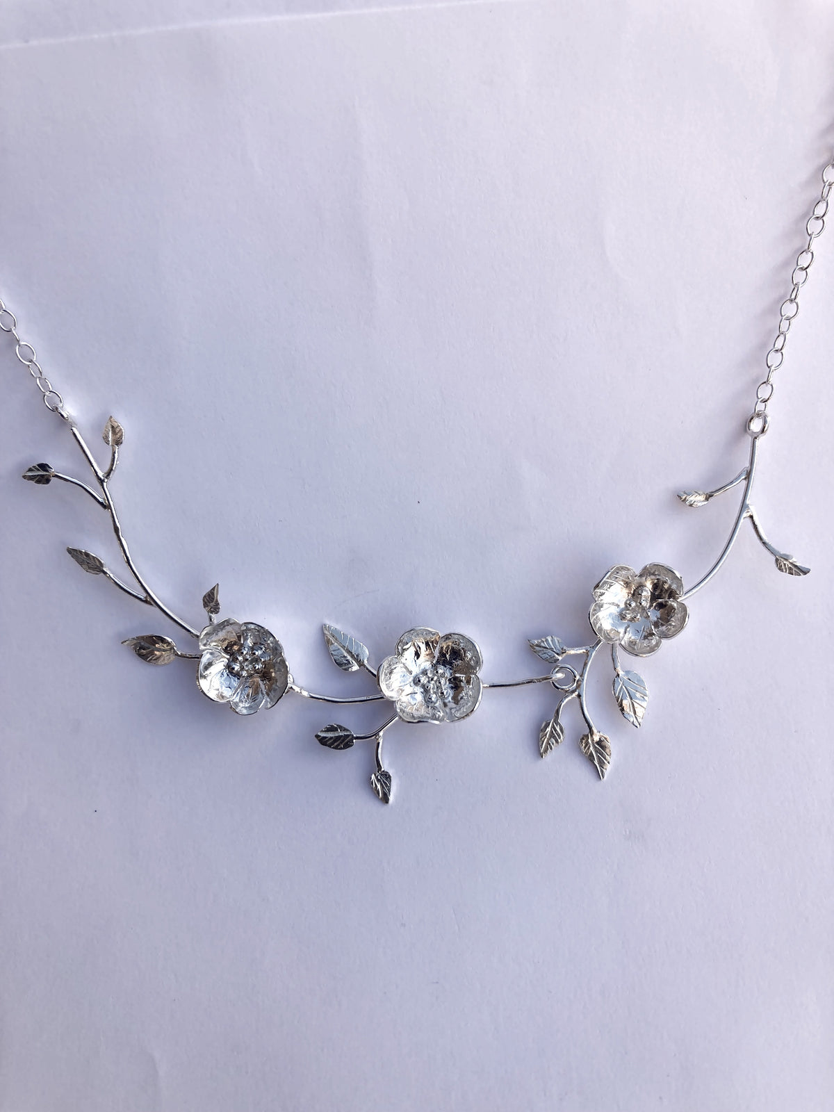 A silver floral necklace by Kim Styles Jewellery featuring three textured wild roses and delicate leaves on a chain, photographed against a plain grey background.