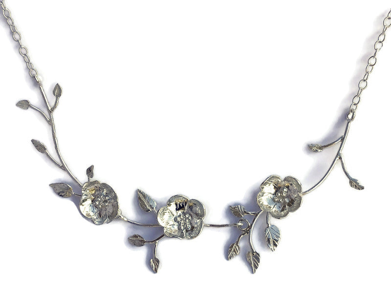 A silver floral necklace by Kim Styles Jewellery featuring three textured flower motifs and delicate leaves on a chain, photographed against a plain white background.