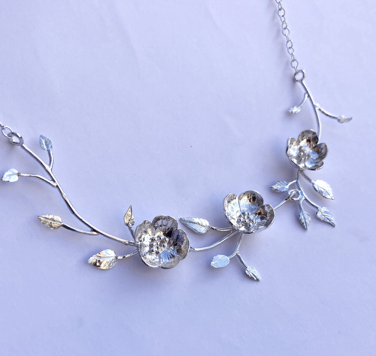 A silver floral necklace by Kim Styles Jewellery featuring three textured wild rose motifs and delicate leaves on a chain, photographed against a plain grey background.