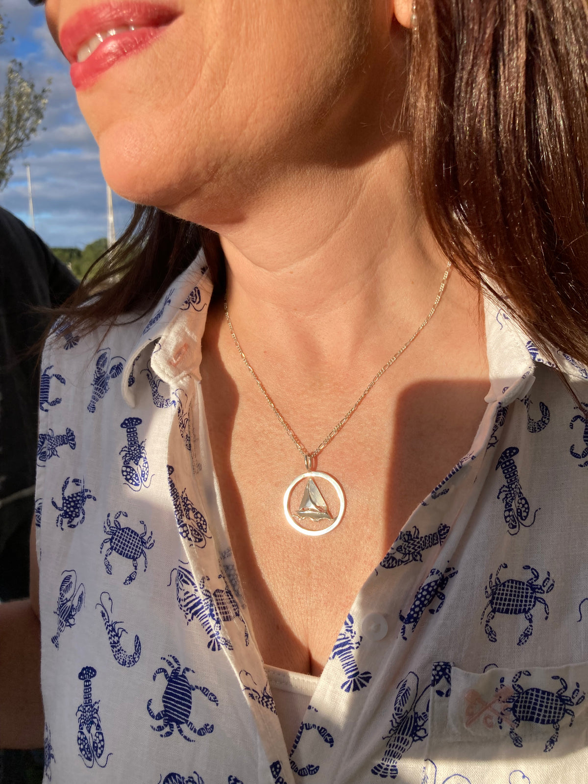 A close-up lifestyle shot of a woman wearing a silver necklace with a circular pendant containing a small 3D sailing boat, paired with a white button-up shirt featuring a blue lobster print.