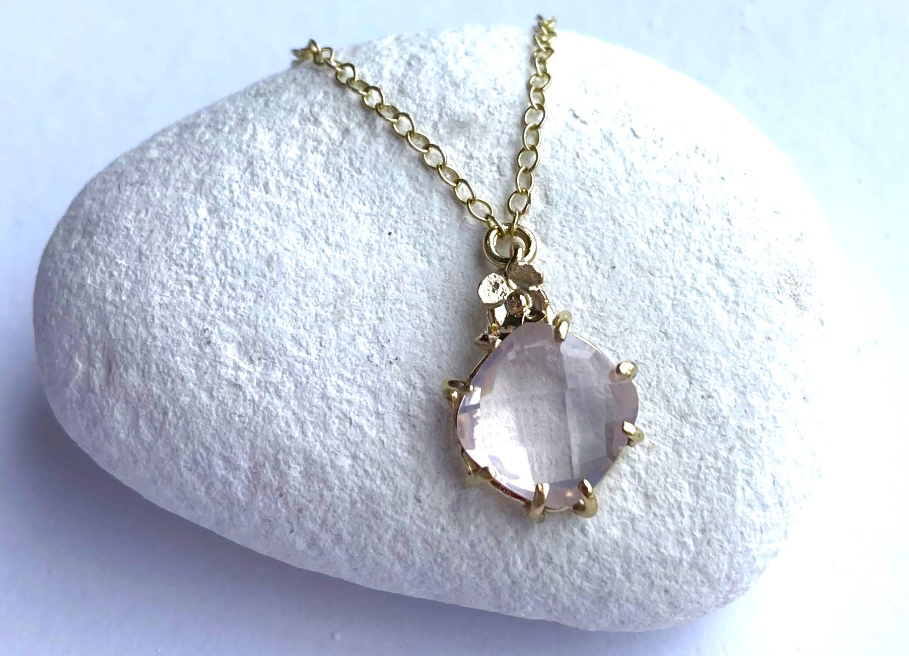 pale pink chequerboard rose quartz  in gold prong setting with tiny flower and gold chain, on white pebble