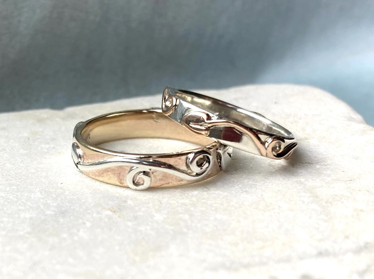 matching fancy wedding rings, one in rose gold with white gold raised scroll details and one in white gold with yellow gold scroll details, resting on marble