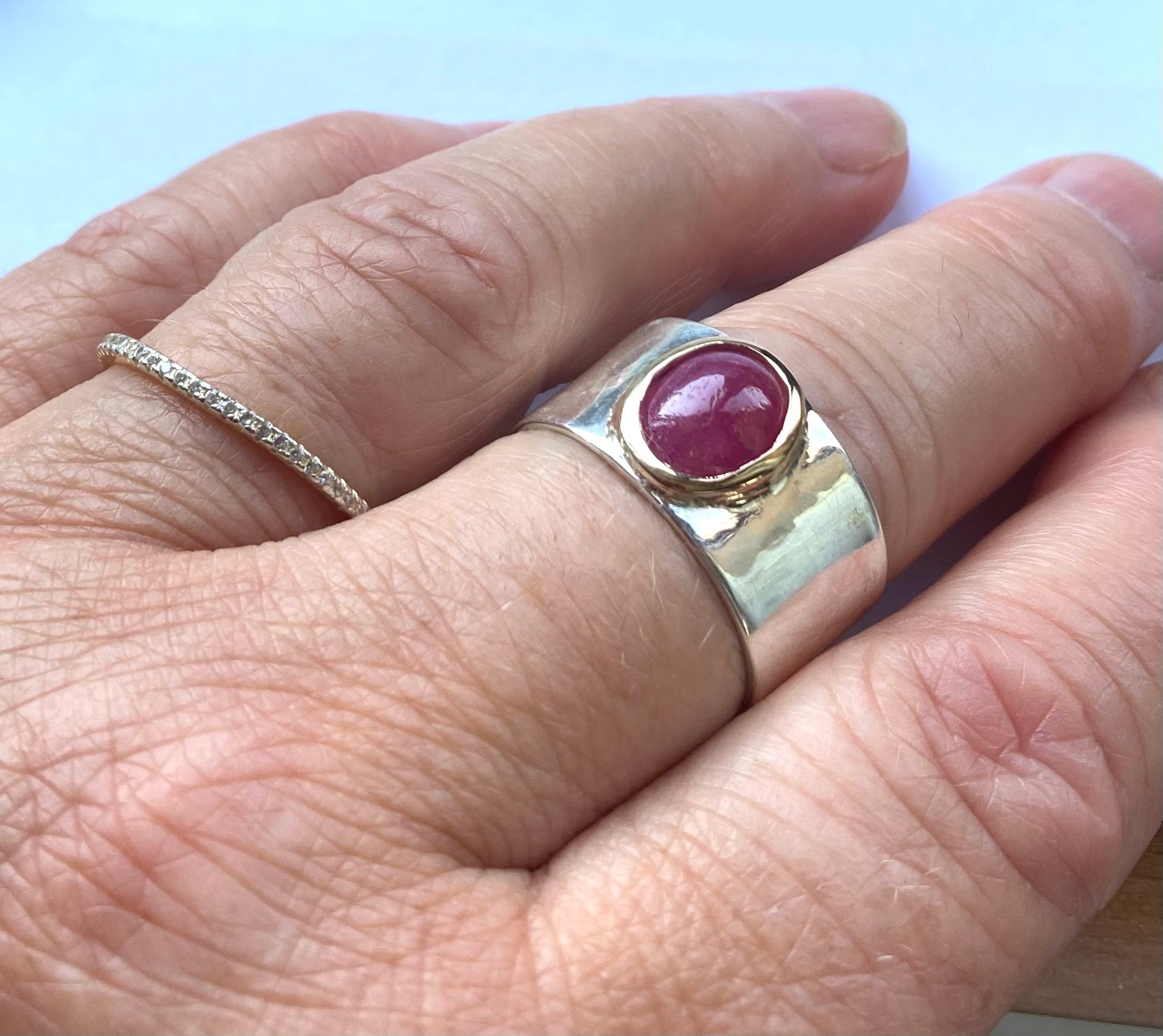 hand with slim diamond ring and wide silver band ring set with oval cabochon ruby with yellow gold setting