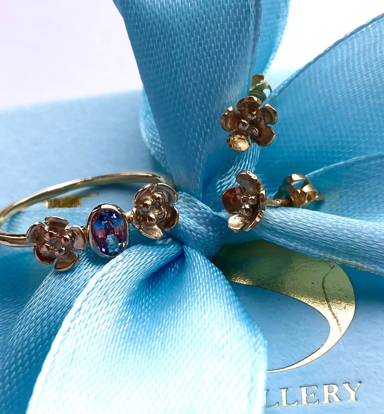 gold floral ring set with blue sapphire and pair of matching gold floral stud earrings, all resting on a pale blue box and  bow