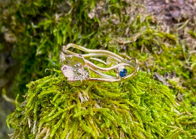 ring of entwined gold wire with a flower and a small round blue sapphire, resting on moss