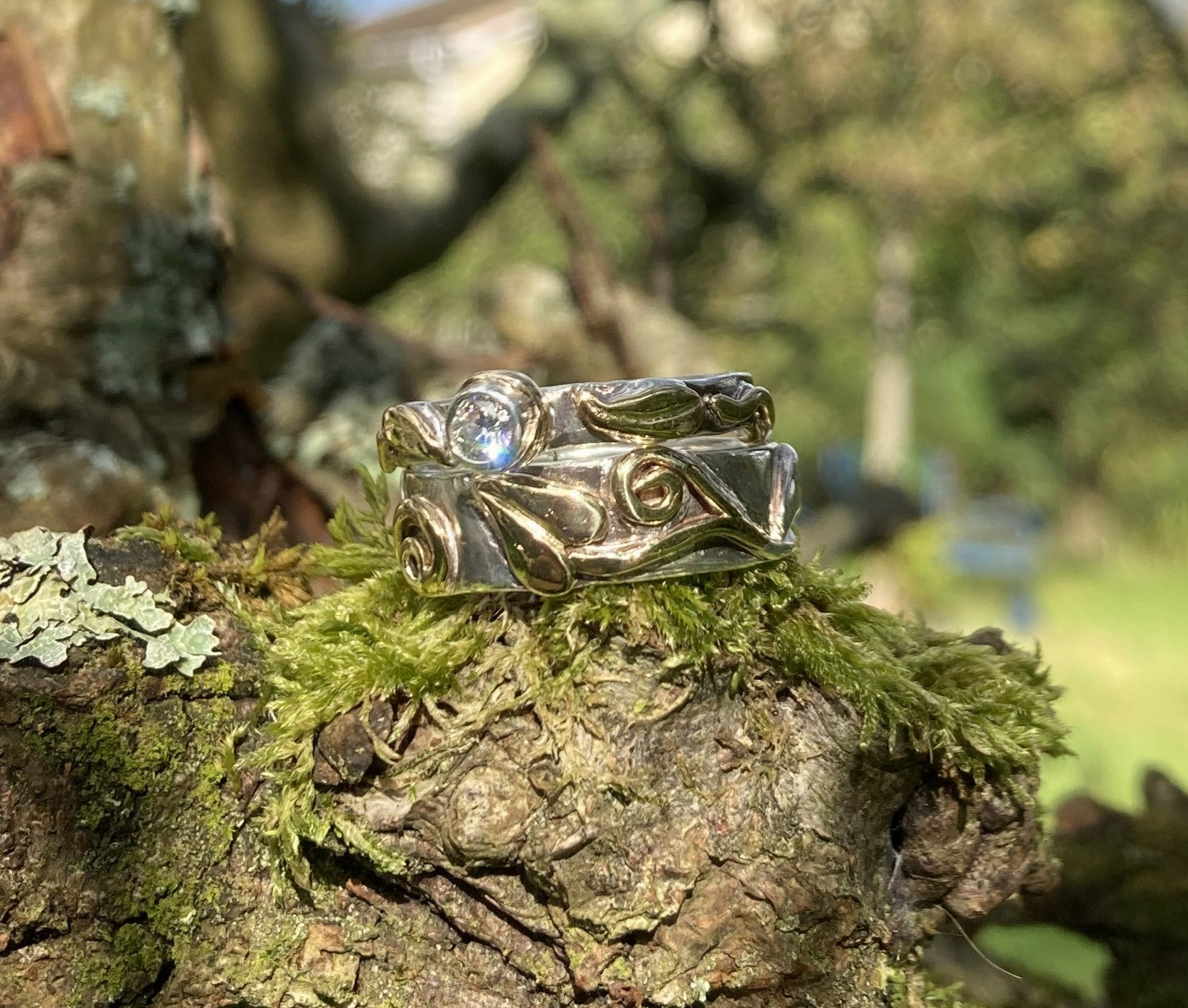 solitaire diamond engagement ring in white gold, with yellow gold floral details in relief, on top of matching white and yellow gold wedding ring, on mossy tree