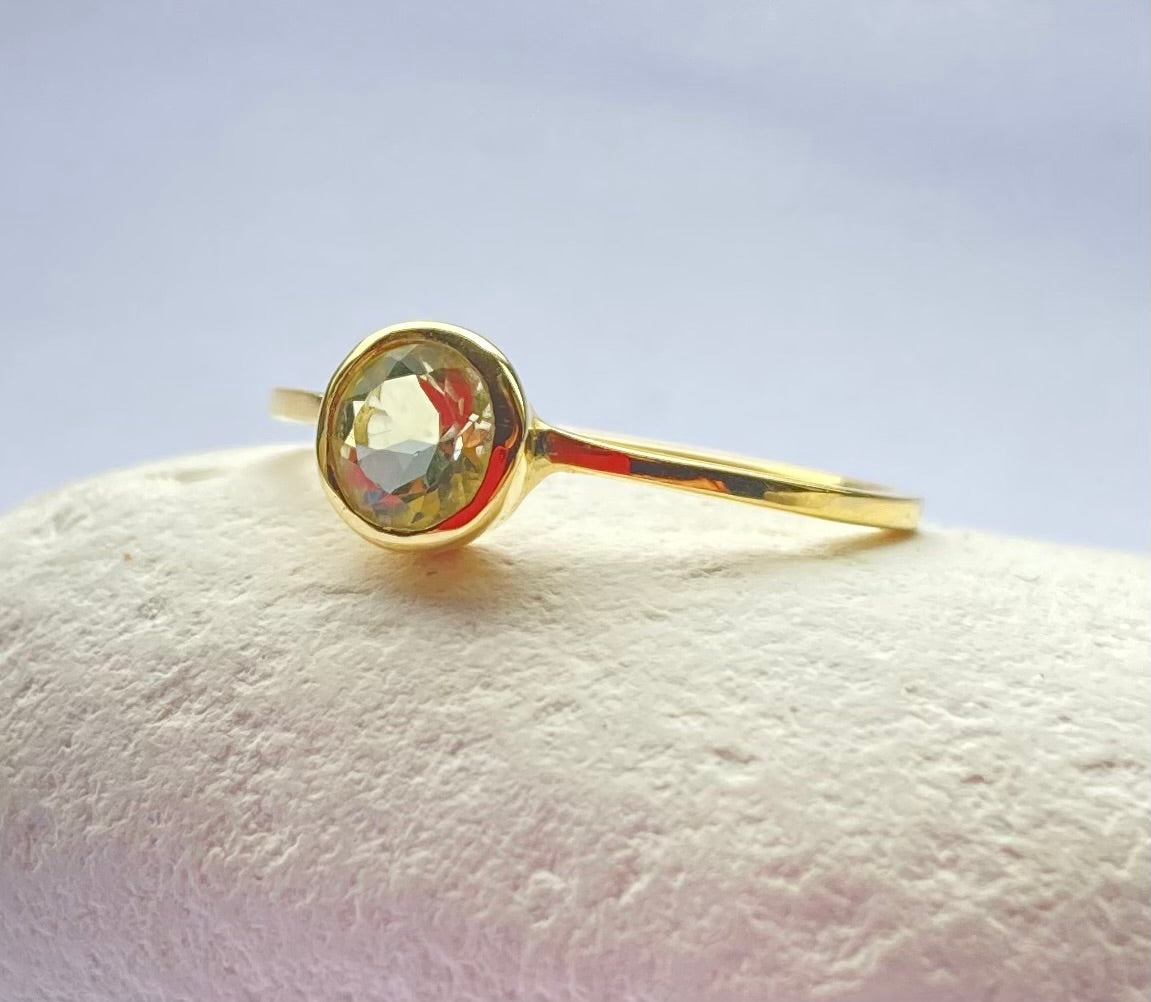 close up of pale green gemstone set in simple slim 18ct yellow gold band ring, resting on white pebble